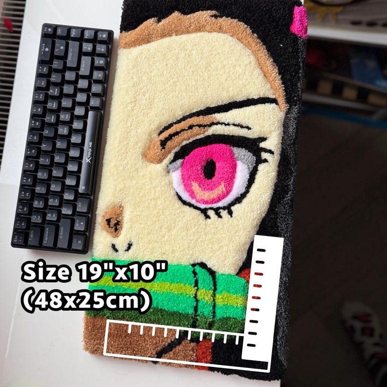 May include: A handmade rug featuring a character's face with a large pink eye, measuring 19" x 10". The rug has a black border and a green and brown scarf. A black keyboard is placed next to the rug.