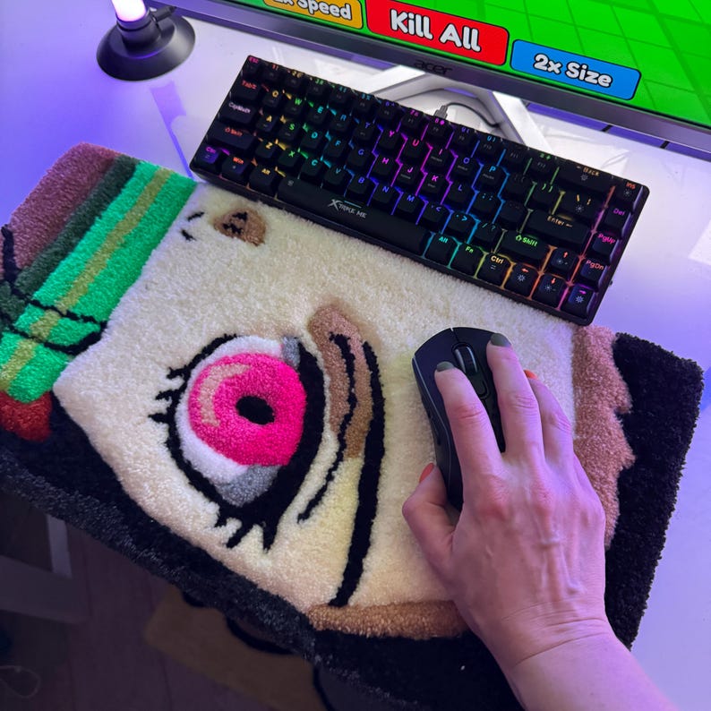 May include: A colorful gaming setup featuring a black keyboard with rainbow backlighting, a black mouse, and a custom rug with a cartoon eye design. The rug has a white background with pink, brown, and black accents.