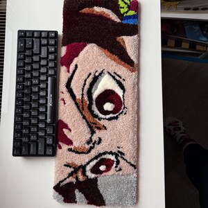 A rectangular, tufted rug featuring a anime face with a large eye. A black keyboard is next to the rug.