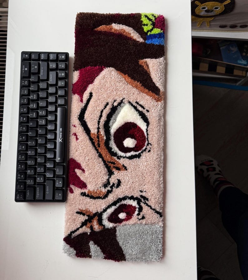 A rectangular, tufted rug featuring a anime face with a large eye. A black keyboard is next to the rug.