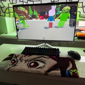 May include: A computer setup with a monitor displaying a video game scene. A black keyboard, mouse, and a rug with an anime eye design are in front of the monitor. The rug is beige, brown, red, and white.