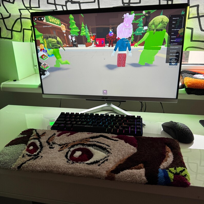 May include: A computer setup with a monitor displaying a video game scene. A black keyboard, mouse, and a rug with an anime eye design are in front of the monitor. The rug is beige, brown, red, and white.