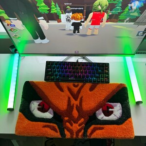 May include: A gaming setup featuring a black keyboard with rainbow-colored keys, resting on a rug with an orange tiger eye design. A computer monitor displays a video game scene. Green and white LED lights are on either side.