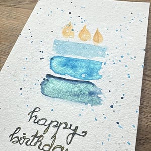 May include: Watercolor birthday card featuring a cake design with blue and teal brushstrokes and gold candle flames. The words "happy birthday" are written in elegant script. The card has a textured, speckled background.