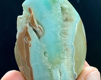 May include: A polished, oval-shaped gemstone with a mix of colors. The stone features a light blue-green hue with bands of brown and tan. The stone is held in a hand, showcasing its natural, organic form. The gemstone has a smooth, polished surface.