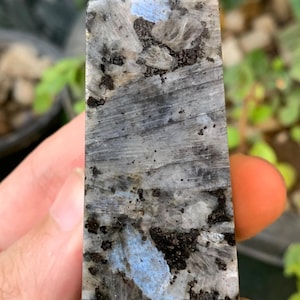 May include: A rectangular crystal point with a flat top. The stone features a mix of grey, black, and blue hues, with a textured surface. The crystal is held in a hand, with a blurred green background.