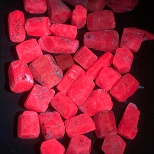 May include: A collection of vibrant, glowing red gemstones. The stones vary in shape, from rectangular to irregular, with a slightly rough texture. The image is set against a dark background, highlighting the stones' intense color.