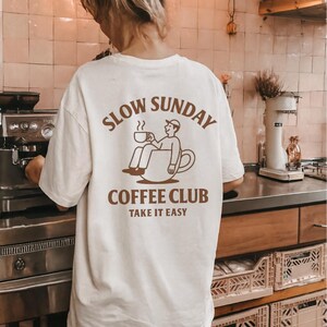以下が含まれることがあります： 「SLOW SUNDAY COFFEE CLUB TAKE IT EASY」と茶色のグラフィックがプリントされたクリーム色のTシャツ。コーヒーカップの中でくつろぐ人物が描かれています。ゆったりとしたオーバーサイズのカジュアルフィットです。