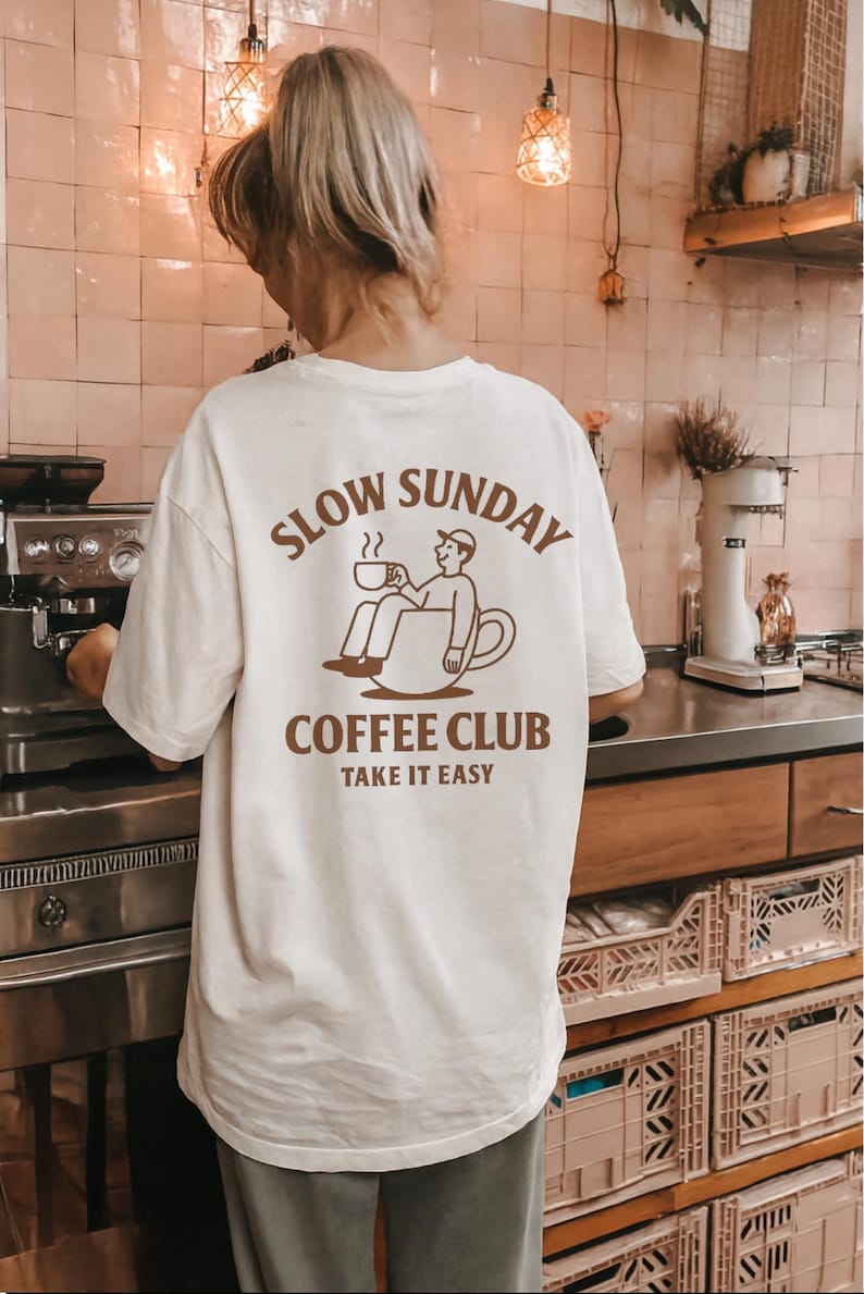 以下が含まれることがあります： 「SLOW SUNDAY COFFEE CLUB TAKE IT EASY」と茶色のグラフィックがプリントされたクリーム色のTシャツ。コーヒーカップの中でくつろぐ人物が描かれています。ゆったりとしたオーバーサイズのカジュアルフィットです。