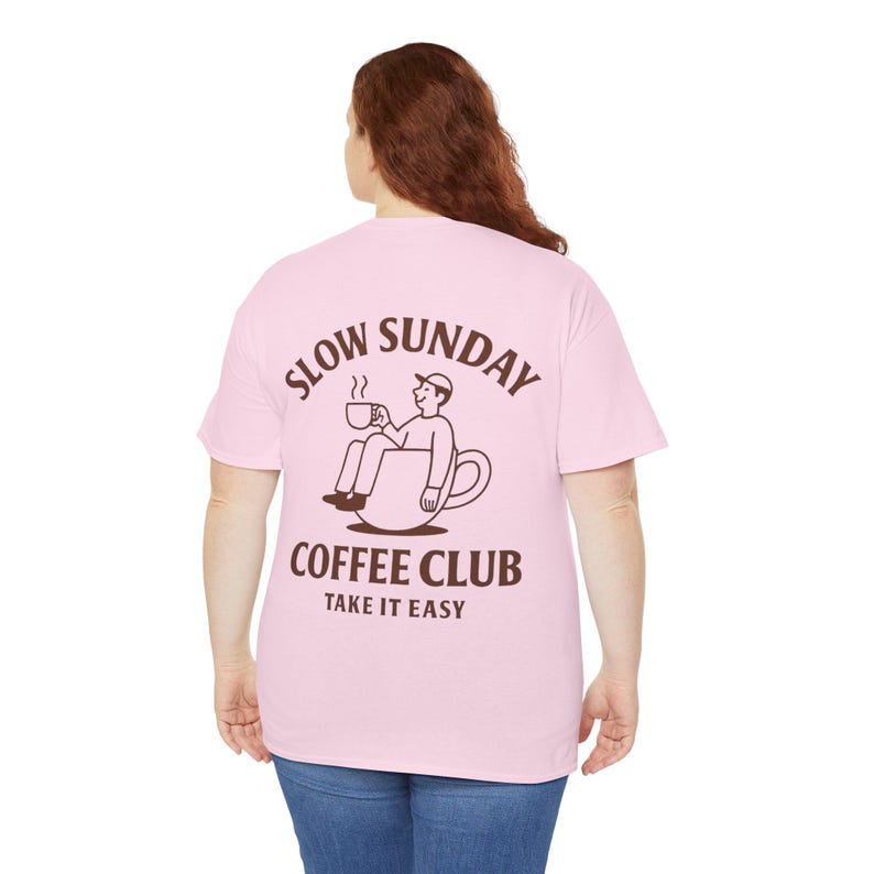 以下が含まれることがあります： 背中に茶色のグラフィックが入った薄いピンク色のTシャツ。グラフィックは、コーヒーカップの中に座ってコーヒーカップを持っている人を特徴とし、茶色の文字で「SLOW SUNDAY COFFEE CLUB TAKE IT EASY」と書かれています。
