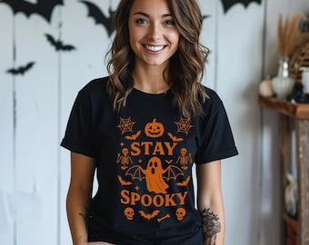 May include: Black t-shirt with orange Halloween-themed graphics, including a pumpkin, ghost, bats, and the words "STAY SPOOKY." The design is centered on the shirt. The model is wearing blue jeans.