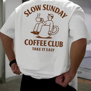 以下が含まれることがあります： 背中に茶色のグラフィックが入った白いTシャツ。グラフィックはコーヒーカップの中に座っている人物で、「SLOW SUNDAY COFFEE CLUB TAKE IT EASY」という茶色の文字が書かれています。