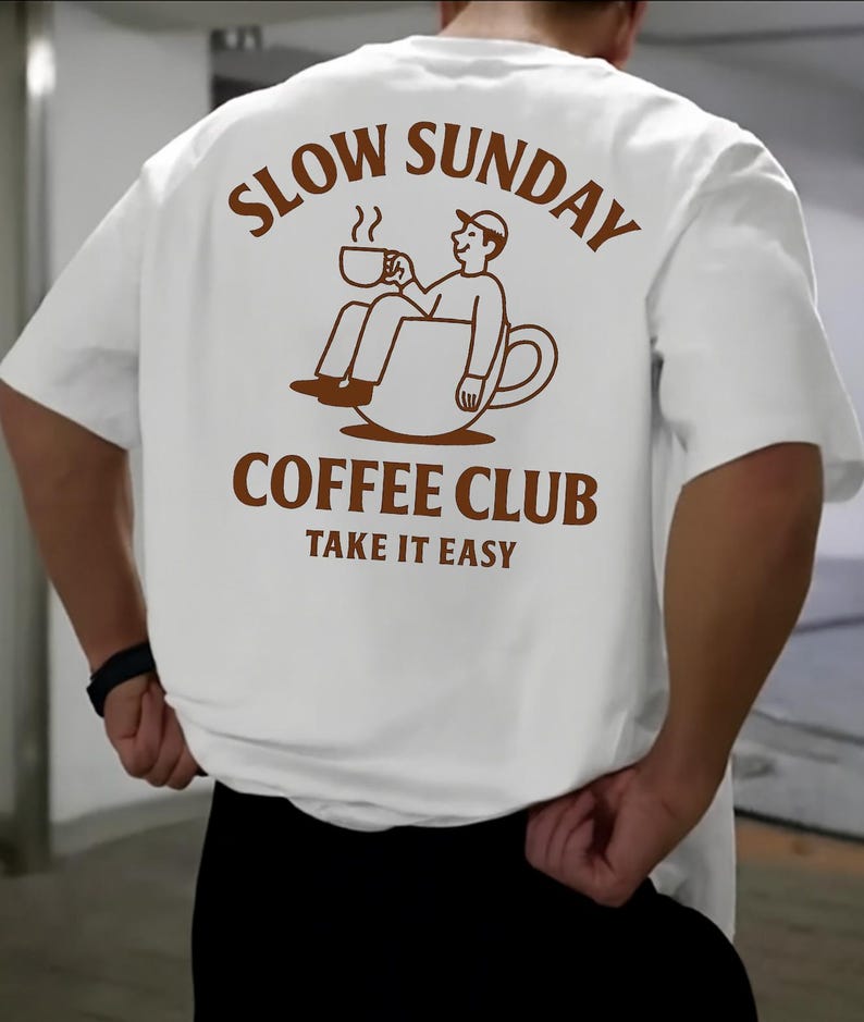 以下が含まれることがあります： 背中に茶色のグラフィックが入った白いTシャツ。グラフィックはコーヒーカップの中に座っている人物で、「SLOW SUNDAY COFFEE CLUB TAKE IT EASY」という茶色の文字が書かれています。