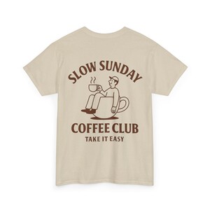 以下が含まれることがあります： ベージュのTシャツ。茶色のグラフィックで、コーヒーカップの中に座ってマグカップを持っている人が描かれています。「SLOW SUNDAY COFFEE CLUB TAKE IT EASY」の文字がグラフィックの上と下にあります。カジュアルウェアです。