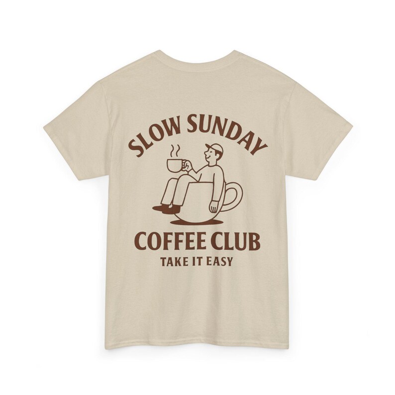 以下が含まれることがあります： ベージュのTシャツ。茶色のグラフィックで、コーヒーカップの中に座ってマグカップを持っている人が描かれています。「SLOW SUNDAY COFFEE CLUB TAKE IT EASY」の文字がグラフィックの上と下にあります。カジュアルウェアです。
