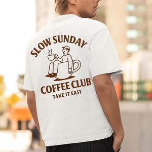 以下が含まれることがあります： 背中に茶色のグラフィックが入った白いTシャツ。「SLOW SUNDAY COFFEE CLUB TAKE IT EASY」と書かれたグラフィックは、コーヒーカップの中に座ってマグカップを持っている人を示しています。