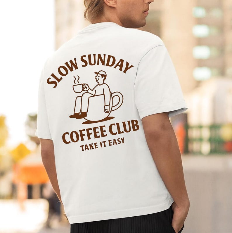 以下が含まれることがあります： 背中に茶色のグラフィックが入った白いTシャツ。「SLOW SUNDAY COFFEE CLUB TAKE IT EASY」と書かれたグラフィックは、コーヒーカップの中に座ってマグカップを持っている人を示しています。