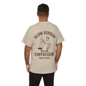 以下が含まれることがあります： 背中に茶色のグラフィックが入ったベージュのTシャツ。グラフィックは、コーヒーカップの中に座っている人物と、茶色の文字で「SLOW SUNDAY COFFEE CLUB TAKE IT EASY」という言葉が特徴です。カジュアルウェアアイテムです。