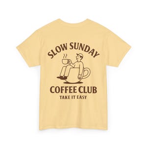 以下が含まれることがあります： 背面に茶色のグラフィックが入った薄い黄色のTシャツ。グラフィックは、コーヒーカップの中に座ってコーヒーカップを持っている人を特徴とし、茶色の文字で「SLOW SUNDAY COFFEE CLUB TAKE IT EASY」と書かれています。