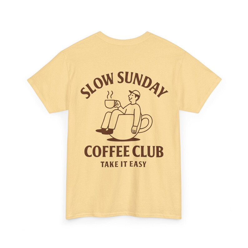 以下が含まれることがあります： 背面に茶色のグラフィックが入った薄い黄色のTシャツ。グラフィックは、コーヒーカップの中に座ってコーヒーカップを持っている人を特徴とし、茶色の文字で「SLOW SUNDAY COFFEE CLUB TAKE IT EASY」と書かれています。