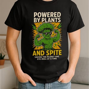 以下が含まれることがあります： 黒いTシャツ。サングラスをかけた筋肉質なブロッコリーのキャラクターのグラフィック。テキストには「POWERED BY PLANTS AND SPITE」と「SAVING THE PLANET ONE EYE-ROLL AT A TIME」と書かれています。