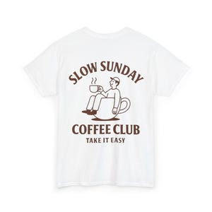 以下が含まれることがあります： コーヒーカップの中に座ってマグカップを持っている人の茶色のグラフィックがプリントされた白いTシャツ。「SLOW SUNDAY COFFEE CLUB TAKE IT EASY」の文字がグラフィックの上と下にプリントされています。