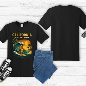Può includere: T-shirt nero con grafica di un surfista e la scritta "CALIFORNIA RIDE THE WAVE". L'immagine include scarpe da ginnastica nere, jeans blu e occhiali da sole. Sfondo in legno bianco.