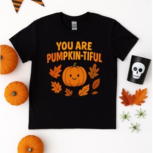 Può includere: T-shirt nera con la scritta arancione "YOU ARE PUMPKIN-TIFUL". La maglietta presenta una grafica di zucca sorridente circondata da foglie arancioni. L'immagine include anche zucche arancioni, una tazza a teschio e decorazioni di Halloween.
