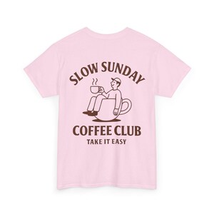 以下が含まれることがあります： 淡いピンク色のTシャツ。茶色のイラストで、コーヒーカップの中に座ってマグカップを持っている人が描かれています。「SLOW SUNDAY COFFEE CLUB TAKE IT EASY」の文字がイラストの上と下にアーチ状に配置されています。
