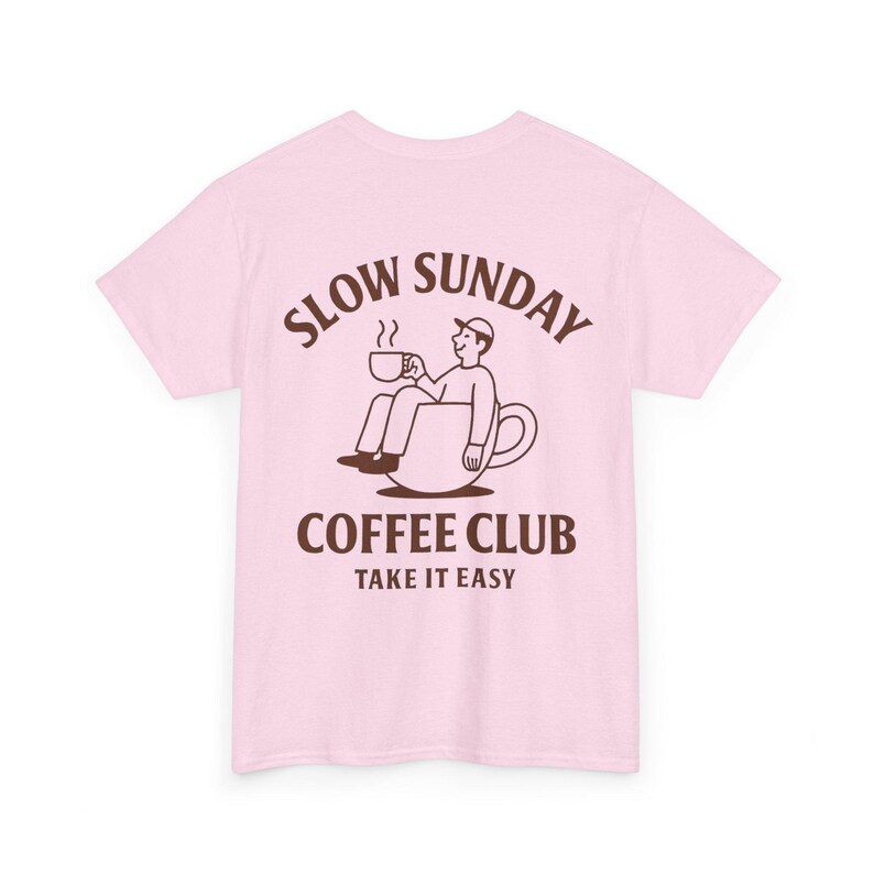 以下が含まれることがあります： 淡いピンク色のTシャツ。茶色のイラストで、コーヒーカップの中に座ってマグカップを持っている人が描かれています。「SLOW SUNDAY COFFEE CLUB TAKE IT EASY」の文字がイラストの上と下にアーチ状に配置されています。