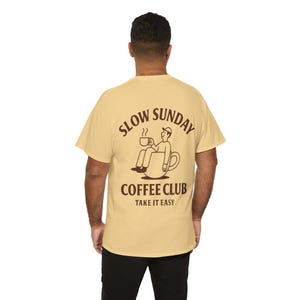 以下が含まれることがあります： 背面には「SLOW SUNDAY COFFEE CLUB TAKE IT EASY」と書かれた、薄いタン色のTシャツ。コーヒーカップの中でくつろぎ、マグカップを持っている人のイラストが描かれています。カジュアルウェアです。
