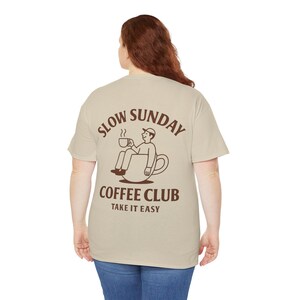 以下が含まれることがあります： 背中に茶色のグラフィックが入ったベージュのTシャツ。グラフィックは、コーヒーカップの中に座ってコーヒーカップを持っている人を特徴とし、茶色のフォントで「SLOW SUNDAY COFFEE CLUB TAKE IT EASY」という言葉が書かれています。