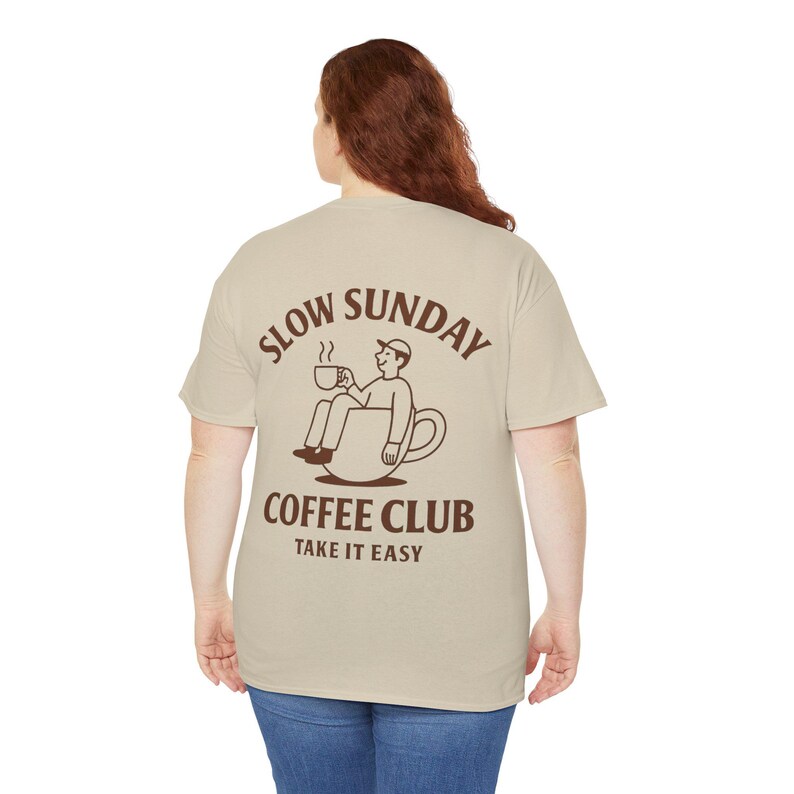 以下が含まれることがあります： 背中に茶色のグラフィックが入ったベージュのTシャツ。グラフィックは、コーヒーカップの中に座ってコーヒーカップを持っている人を特徴とし、茶色のフォントで「SLOW SUNDAY COFFEE CLUB TAKE IT EASY」という言葉が書かれています。
