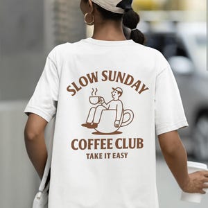 以下が含まれることがあります： 背中に茶色のグラフィックが入った白いTシャツ。「SLOW SUNDAY COFFEE CLUB」の文字と、コーヒーカップでくつろぐ人のイラストが描かれています。「TAKE IT EASY」の文字が下にあります。