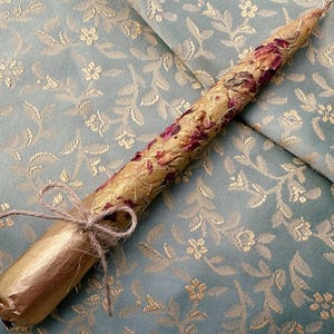May include: A decorative candle with a gold base and a floral design. The candle is wrapped with dried rose petals and tied with twine. The background is a light blue fabric with a floral pattern.