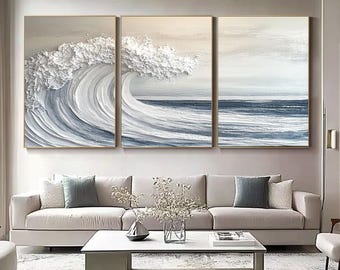 May include: Three-panel wall art depicting a textured ocean wave in shades of white and blue. The artwork is framed in gold and hangs above a beige sofa with decorative pillows. The overall theme is coastal decor.