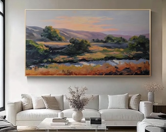 May include: Large landscape oil painting featuring a sunset over a field and mountains. The artwork uses a palette of orange, green, and blue hues. The painting is framed in a gold-colored frame and hangs above a white sofa.