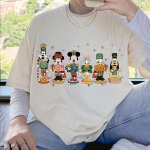 May include: Cream-coloured t-shirt featuring a festive design of Disney characters as nutcrackers. The characters include Goofy, Minnie Mouse, Mickey Mouse, Daisy Duck, Donald Duck, and Pluto, each in a unique uniform. A string of lights is at the bottom.