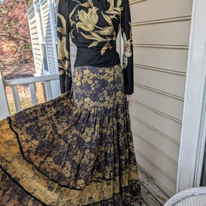 Puede incluir: Un conjunto de top cruzado negro y falda larga. El top presenta un estampado floral en dorado y crema, con mangas largas. La falda es una falda larga escalonada con un diseño floral similar y una banda dorada en el dobladillo. El conjunto se muestra en un maniquí.