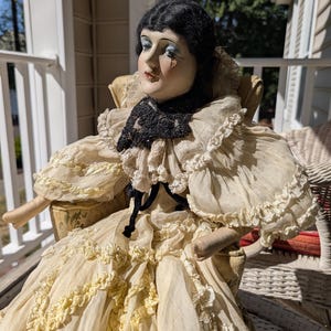 May include: A vintage porcelain doll with a painted face, black hair, and a cream-colored dress with ruffled details. The doll is seated in a chair and wears a black lace collar.
