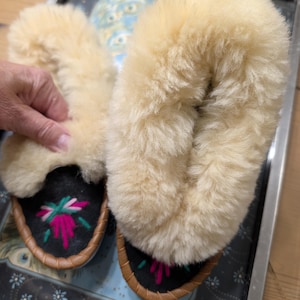 May include: A pair of cream-colored, fluffy slippers with black soles and brown leather trim. The soles feature a floral embroidery design in pink, green, and white. The slippers are displayed on a decorative tray.