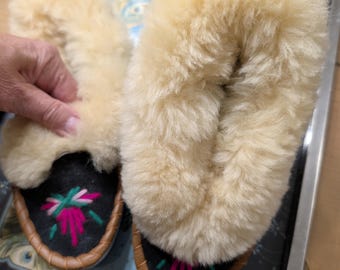 Könnte beinhalten: Ein Paar cremefarbene, flauschige Hausschuhe mit schwarzen Sohlen und braunen Ledereinfassungen. Die Sohlen weisen ein Blumenstickerei-Design in Rosa, Grün und Weiß auf. Die Hausschuhe werden auf einem dekorativen Tablett präsentiert.
