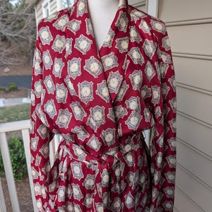 May include: A burgundy-colored robe with a repeating clock face pattern in cream and gray. The robe has long sleeves, a wrap-around design, and a matching belt. The robe is displayed on a mannequin.