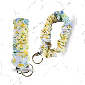 May include: Two floral fabric keychains. One is a wristlet style, and the other is a long, looped keychain. Both are made of a yellow fabric with a pattern of small yellow flowers and blue leaves. Each keychain has a silver-colored metal ring.