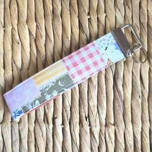 May include: A fabric keychain featuring a patchwork design with sections of red and white gingham, light purple, and yellow patterns. The rectangular fabric strap is attached to a silver-toned metal hardware clamp with a circular split ring.