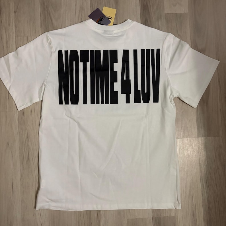 May include: White t-shirt with the text "NO TIME 4 LUV" printed in large, bold, black letters across the back. The shirt has short sleeves and a tag attached at the neckline. The shirt is displayed on a light wood surface.