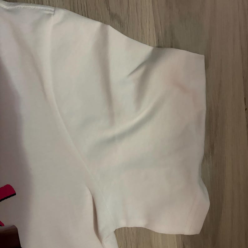 May include: Close-up of a white t-shirt sleeve. The fabric is smooth and appears to be made of a lightweight material. The sleeve is slightly wrinkled, suggesting it has been worn or folded. The background is a light-colored wooden surface.