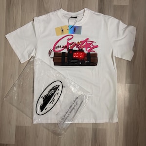 May include: White t-shirt with a graphic of a bomb with the word "Crazy" in pink script. The bomb has a digital display showing "00:01". A clear plastic bag with a logo and the quote "The life you live is either a daring adventure, or nothing at all" is next to the shirt.