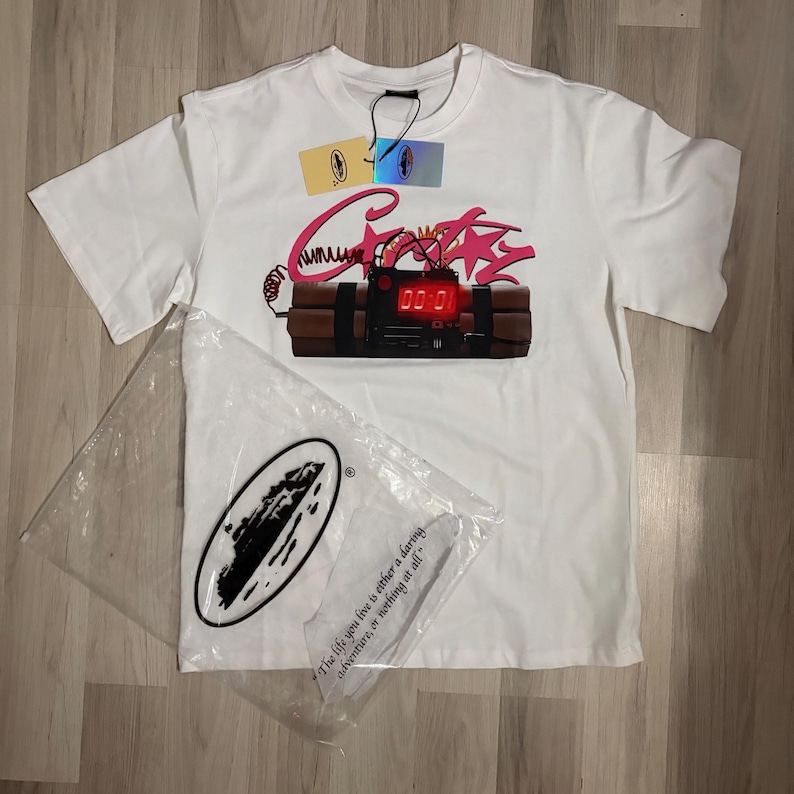 May include: White t-shirt with a graphic of a bomb with the word "Crazy" in pink script. The bomb has a digital display showing "00:01". A clear plastic bag with a logo and the quote "The life you live is either a daring adventure, or nothing at all" is next to the shirt.