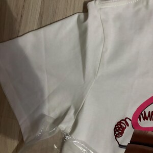 May include: White t-shirt with a graphic design. The design features a cartoon image with pink and brown accents. The shirt is folded and partially covered in a clear plastic bag.